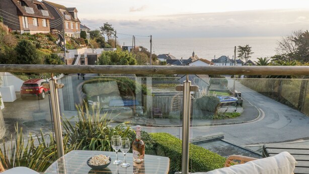 A balcony with a glass table, wicker chairs, and a view of the sea and houses. - A Stone's Throw (Torpoint)