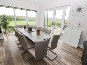 A modern dining room with a large table, comfortable chairs, and a view of the countryside through large windows. - Ty Marlyn (Swansea)