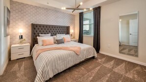 Een slaapkamer met een gestoffeerd hoofdeinde, een groot bed met gestreepte beddengoed, een nachtkastje met een lamp en een grote spiegel aan de muur.
