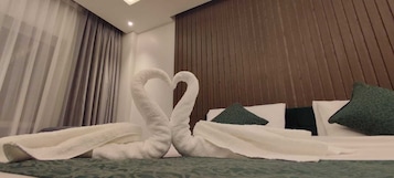 A hotel room with a bed featuring a heart-shaped towel arrangement, green patterned bedspread, and dark wooden headboard.
