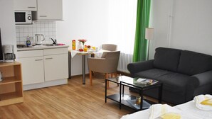 Habitación de hotel con cama, sofá, mesa de comedor, zona de cocina y ventana con cortinas.