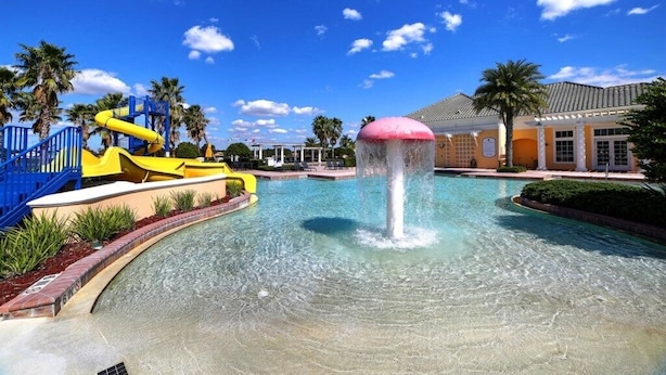 A swimming pool with a water slide and a fountain, surrounded by palm trees and a building with a tiled roof. - Providence Golf & Country Club Executive Pool Home - Located Near Disney (Davenport)