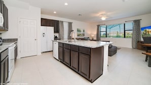 A spacious kitchen with dark cabinetry, a large island, and a view of a green landscape through large windows. - Providence Golf & Country Club Executive Pool Home - Located Near Disney (Davenport)