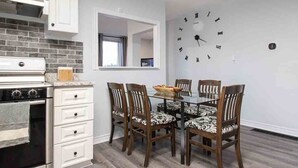 A modern kitchen with white cabinets and a black stove, a dining area with a glass table and patterned chairs, and a wall clock.