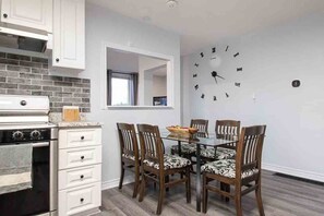 Una cocina moderna con gabinetes blancos y una estufa negra, un área de comedor con una mesa de vidrio y sillas estampadas, y un reloj de pared.