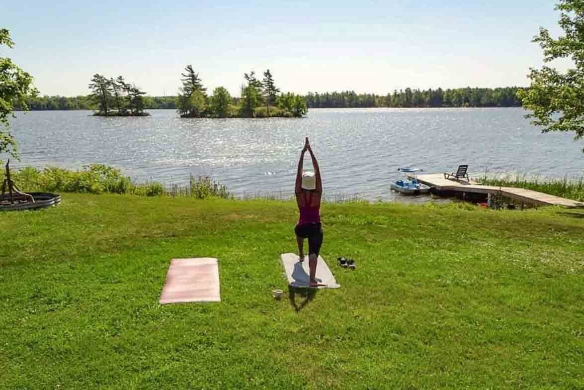 Une personne effectue une posture de yoga sur un tapis, près d’un lac, avec des bateaux et une jetée en arrière-plan.