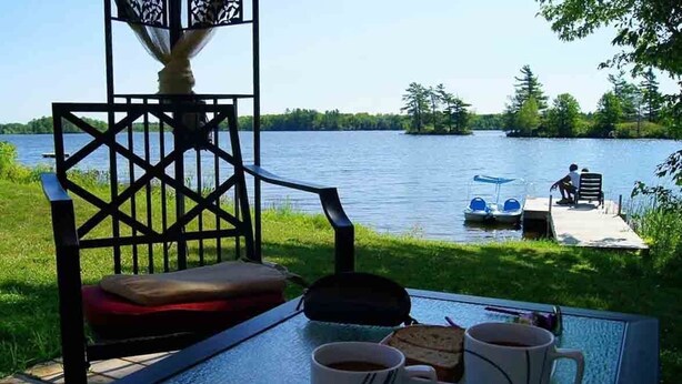 A glass table with two mugs and a sandwich, a chair, and a view of a lake with a dock and a boat.