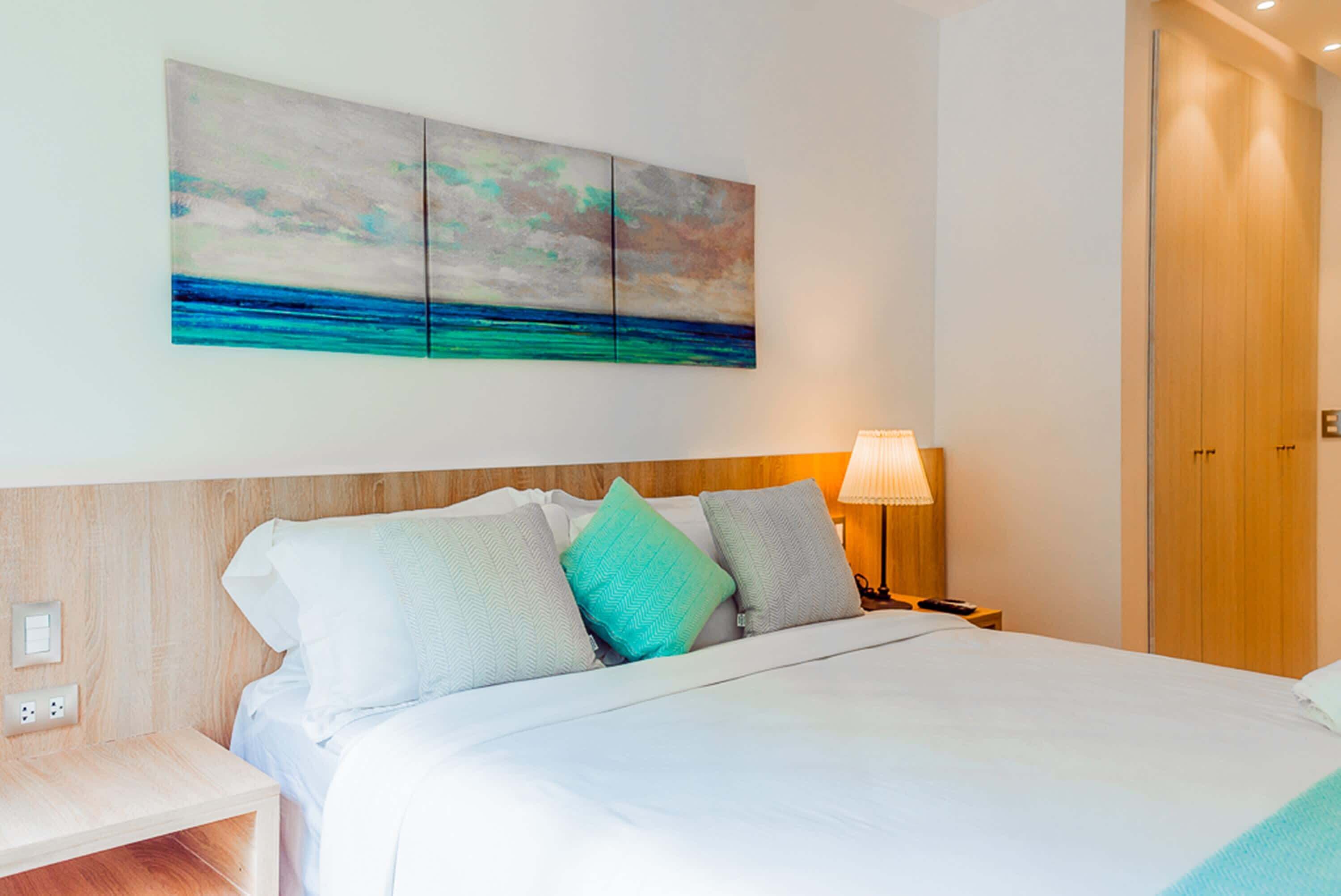 Una habitación de hotel con una cama, una mesita de noche, una lámpara y un cuadro de una playa.