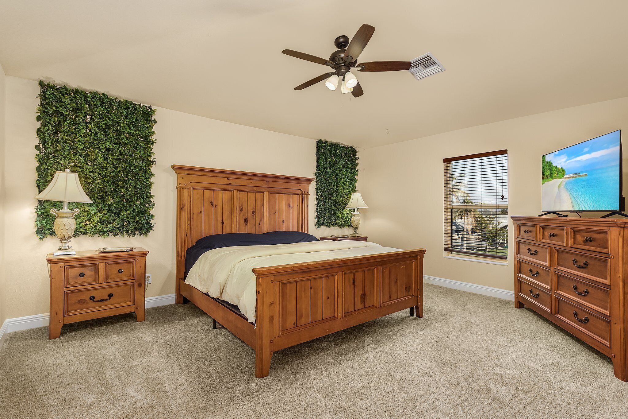 Une chambre à coucher avec un lit en bois, deux tables de chevet, une commode, un ventilateur de plafond, un téléviseur fixé au mur et une fenêtre avec des stores.