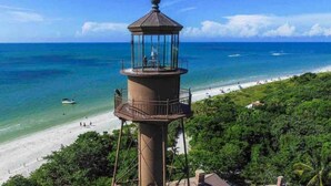 A lighthouse with a glass enclosure, situated on a cliff overlooking a beach with clear blue waters and a sandy shore.