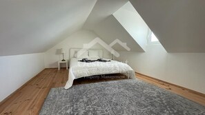Un dormitorio con techo inclinado, una cama con colcha blanca, una mesita de noche pequeña con lámpara y una alfombra gris estampada.