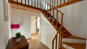Un escalier avec des rampes en bois mène à l’étage supérieur. Une table en bois avec une plante en pot est visible au premier plan.
