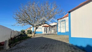 Un chemin pavé mène à un bâtiment aux murs bleus et blancs, doté d’un toit en tuiles et d’un arbre en premier plan.