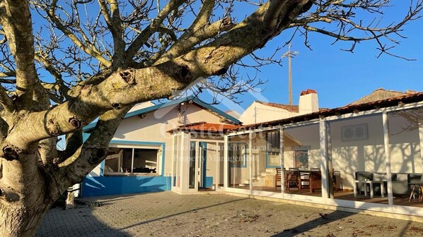 Una propiedad con un gran árbol en primer plano, un área de estar cubierta al aire libre y un edificio con puerta azul.