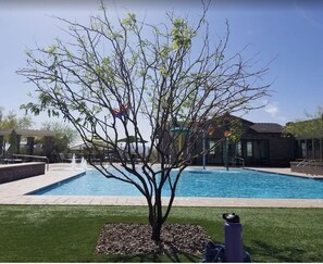 Ein Swimmingpool mit einem Baum im Vordergrund, einer Terrassenfläche und einem Gebäude im Hintergrund.
