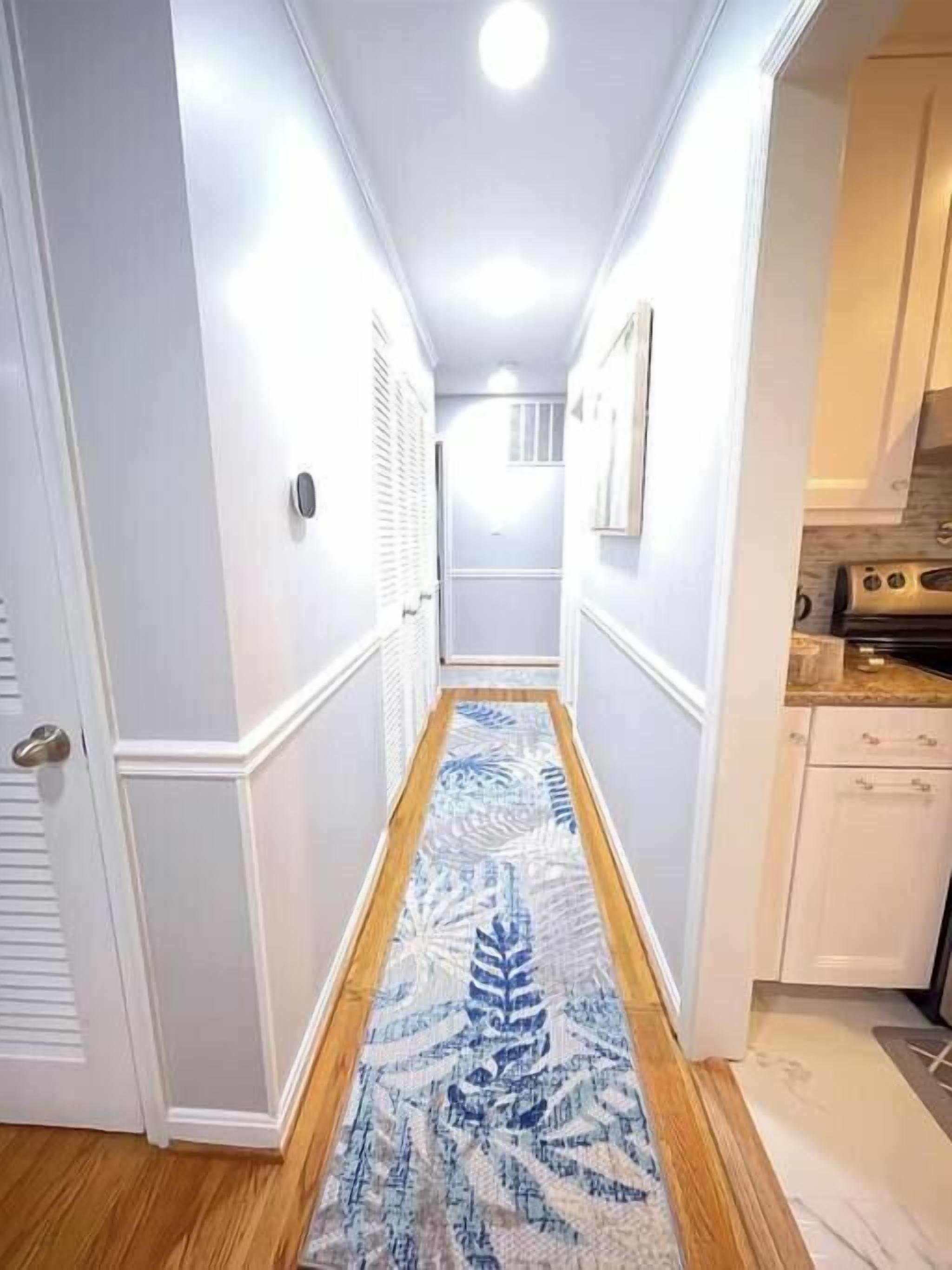 Un couloir étroit, orné d’un tapis à motifs, avec des murs blancs et un coin cuisine comprenant des placards et une cuisinière.