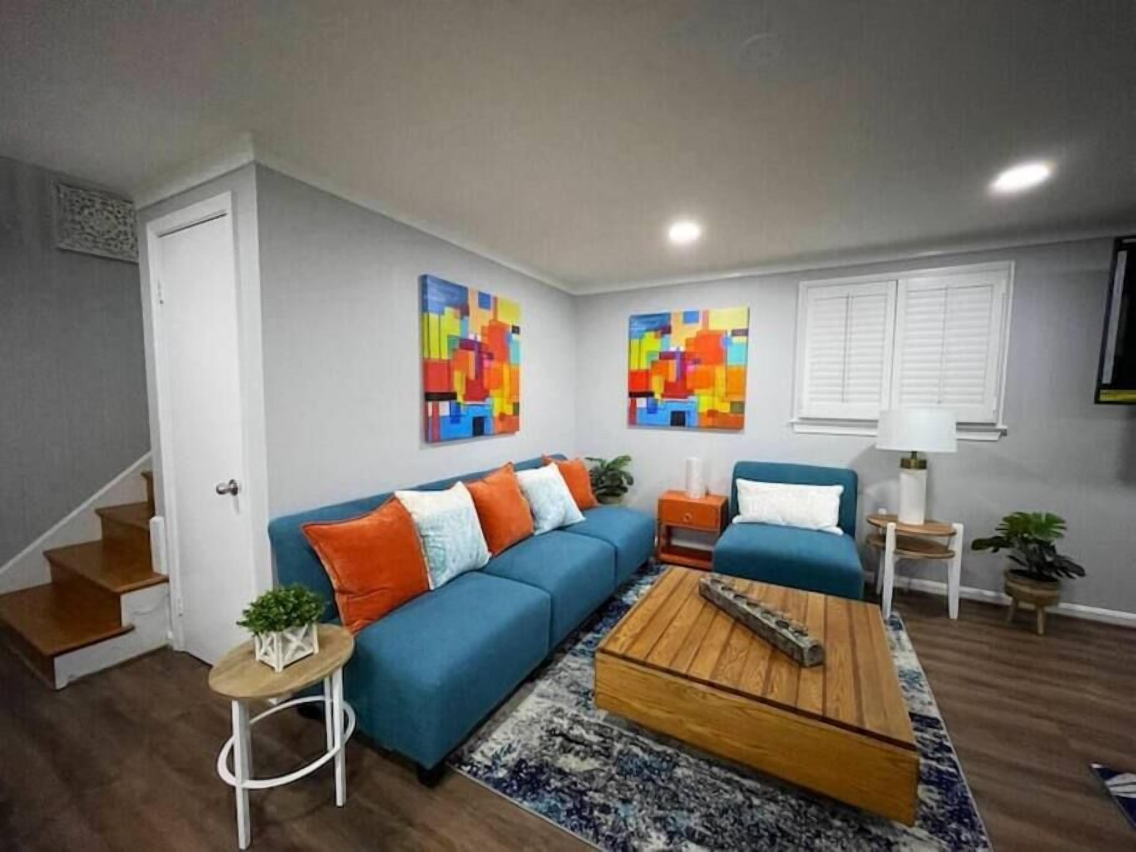 Un salon moderne avec un canapé bleu, des tableaux abstraits aux couleurs vives, une table basse en bois et un escalier.