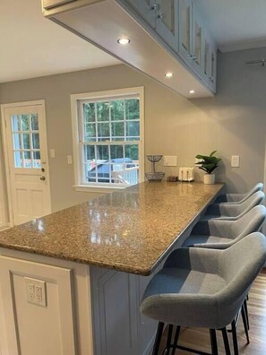 A kitchen with a granite countertop, grey bar stools, and a window with a view of trees.