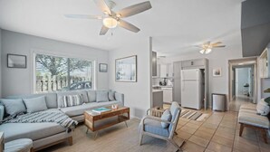Un salon moderne comprenant un canapé, une table basse et un fauteuil, un coin cuisine avec des placards et des appareils électroménagers, et un ventilateur de plafond.