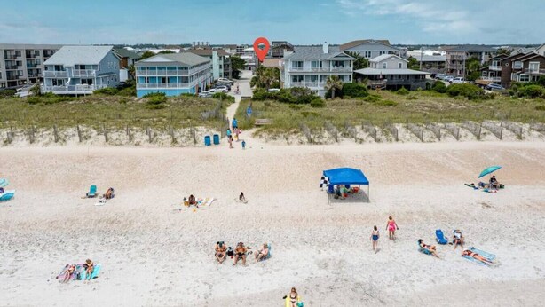 Une plage où des gens se prélassent au soleil et se baignent, une tente bleue et des maisons en arrière-plan.