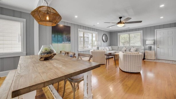 A spacious living room with a wooden dining table, a ceiling fan, and a large window with blinds. - A Swell Life | Walking Distance to Ocean w/ King B (Wrightsville Beach)