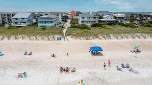 A beach with people sunbathing and swimming, a blue tent, and houses in the background. - A Swell Life | Walking Distance to Ocean w/ King B (Wrightsville Beach)