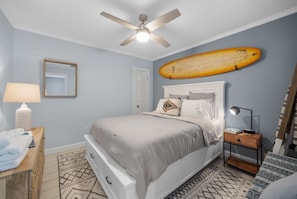 Un dormitorio con una cama, ventilador de techo, una tabla de surf, mesita de noche, lámpara y espejo.