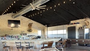 A spacious room with a large table, chairs, a mounted deer head, and a stone wall with a mounted television. - Bunkhouse in Texas ranch country (Guthrie)
