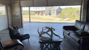 A garage with a concrete floor, a stone wall, a black leather couch, a polka-dotted chair, and a wooden coffee table with antlers. - Bunkhouse in Texas ranch country (Guthrie)