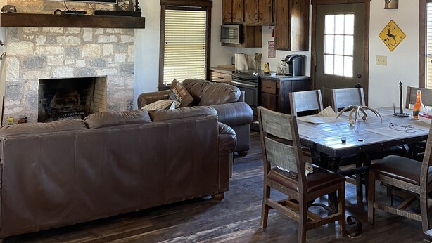 A spacious living area with a stone fireplace, a large leather sofa, wooden dining table, and a kitchen in the background. - Bunkhouse in Texas ranch country (Guthrie)