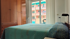 Een slaapkamer met een bed, een kledingkast, een raam met uitzicht op gebouwen en een vloerlamp.