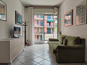 A living room with a green sofa, a wooden coat rack, and a view of colorful buildings through a glass door. - Colombo 7 by Tempovacanza (Marina di Andora)