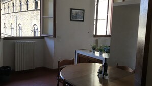 A dining area with a round table, chairs, a radiator, a window with a view of a stone building, and a framed picture on the wall.