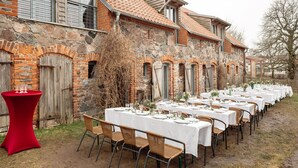An outdoor dining area with white tables and wicker chairs, set against a stone building with a tiled roof. - Stylishly furnished fieldstone barn with dining room and terrace by the village pond (Werneuchen)