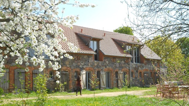 A stone building with a tiled roof, arched windows, and a pathway leading to it. - Stylishly furnished fieldstone barn with dining room and terrace by the village pond (Werneuchen)