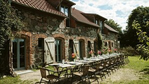 A stone building with a tiled roof, outdoor dining area with tables and chairs, and a grassy lawn. - Stylishly furnished fieldstone barn with dining room and terrace by the village pond (Werneuchen)