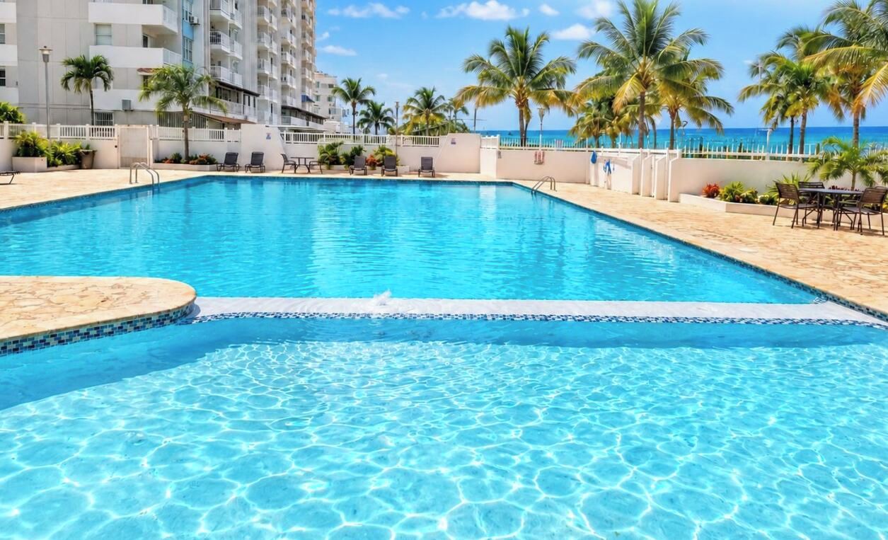 A clear swimming pool with a white deck, surrounded by palm trees and a building in the background.