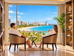 Ein Zimmer mit großem Fenster mit Blick auf Strand und Stadtansicht, zwei sich gegenüberliegende Stühle, ein kleiner Tisch mit einer Vase und ein Bücherregal mit dekorativen Gegenständen.