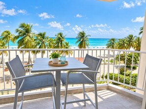 A balcony with a table and chairs, overlooking a beach with palm trees and the ocean.