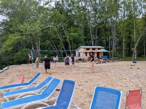 A beach volleyball court with blue and red lounge chairs, a wooden cabin, and dense forest in the background.