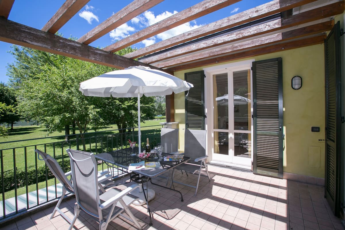 A covered patio with a table and chairs, an umbrella, and a view of a green park.