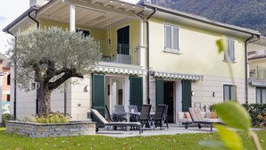 A two-story house with a balcony, outdoor seating, and a tree in the garden. - Villa Apartments - Porto Letizia (Porlezza)