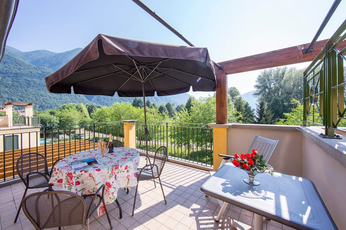 An outdoor terrace with a table and chairs, a large umbrella, and a view of mountains.