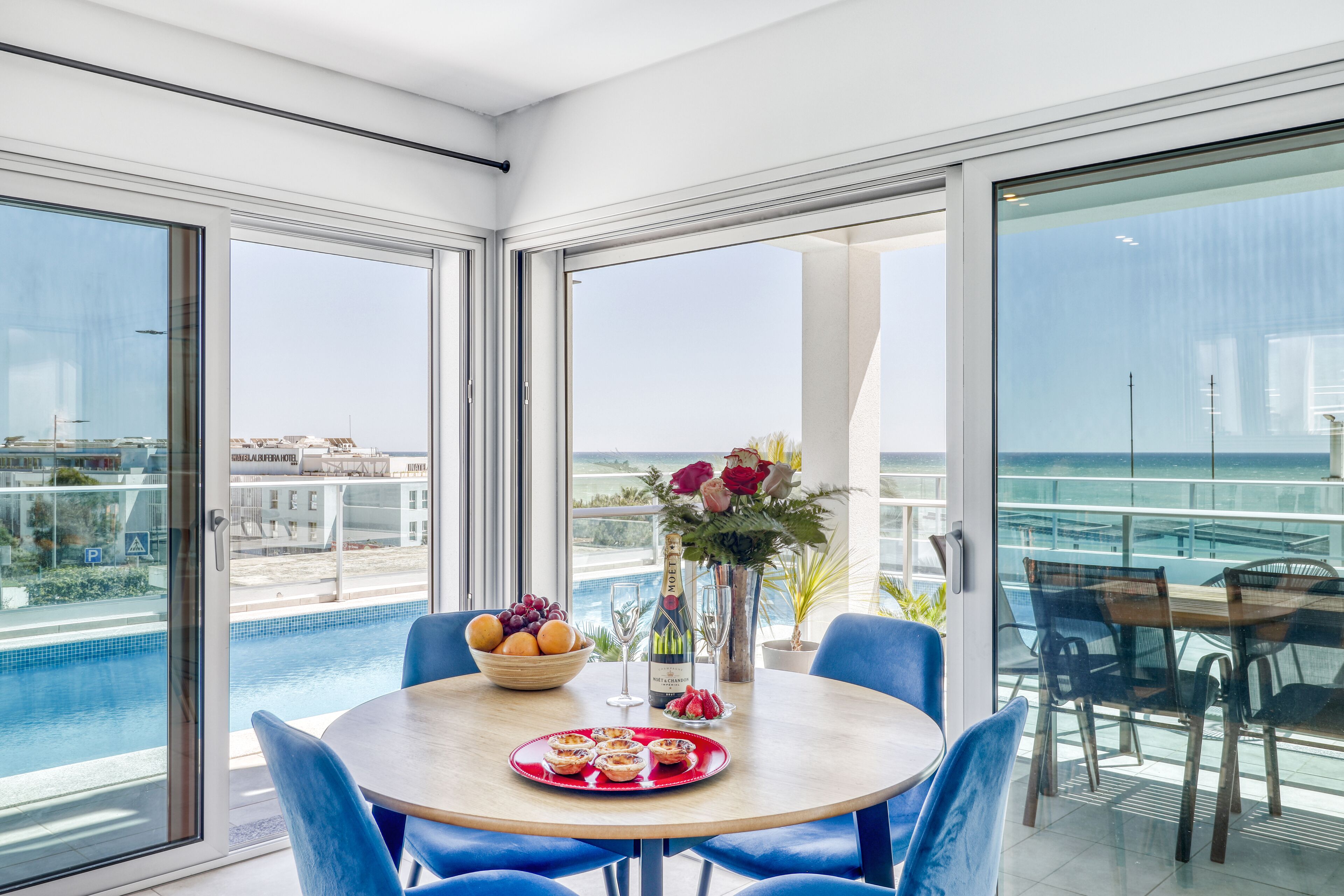A dining area with a table set for two, a bowl of fruit, a bottle of champagne, and a vase of flowers.