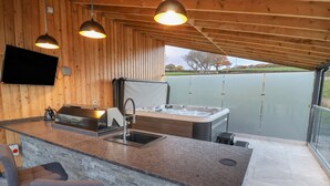 A modern kitchen with a built-in stove, sink, and a large island. There is a bar area with stools and a TV mounted on the wall. - Fron Haul - Gwennol (Ruthin)