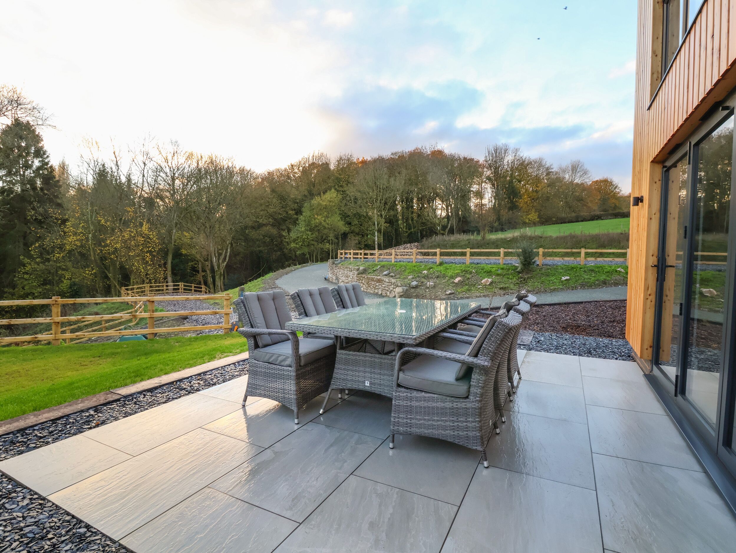A patio with a glass-top table and wicker chairs, surrounded by a stone wall and a wooden fence.