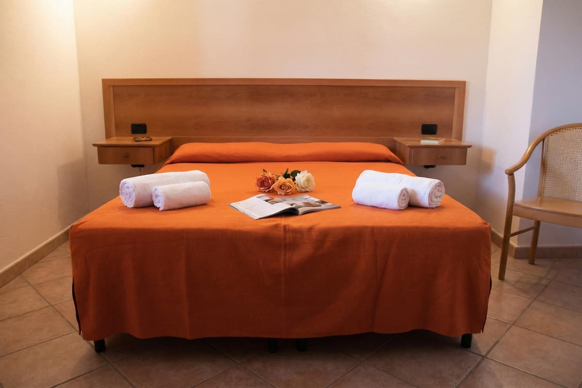 Uma cama bem arrumada com colcha laranja, duas mesas de cabeceira com abajures, uma cabeceira de madeira e uma única cadeira.