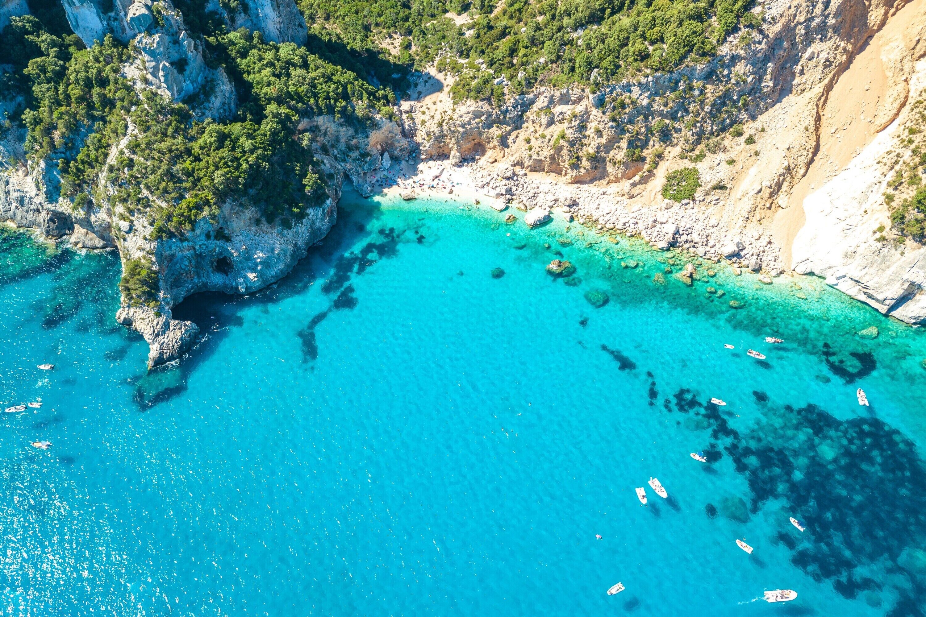 An aerial view of a coastal area with clear turquoise waters, rocky cliffs, and sparse vegetation.