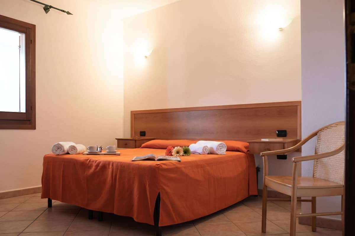 Cama bem arrumada com cabeceira de madeira, uma cadeira e uma mesa pequena com uma xícara e um livro.