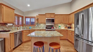 A kitchen with wooden cabinets, a stainless steel refrigerator, granite countertops, and a central island with stools. - Amenities Galore! Outdoor Living & Lots of Games (Peoria)